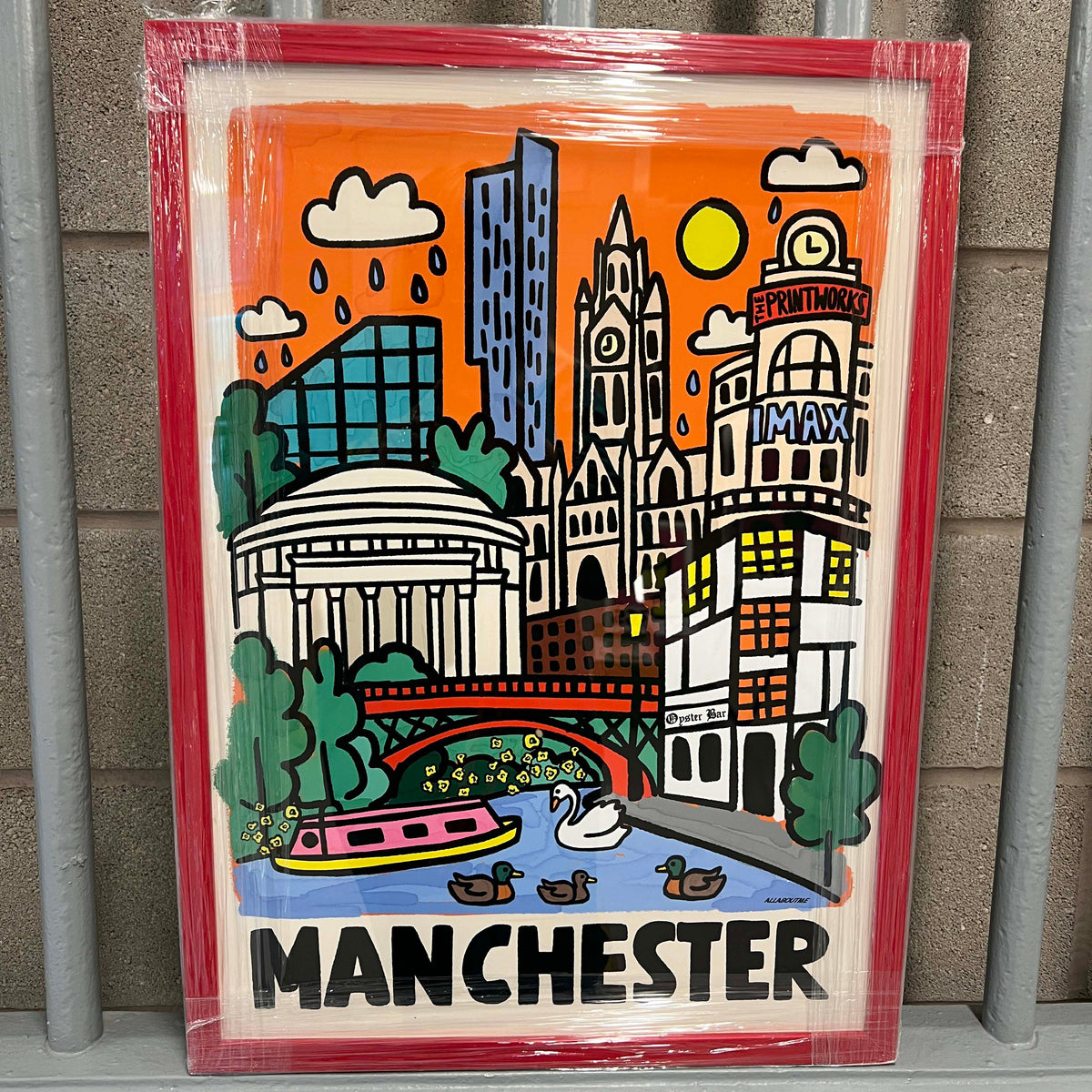 A framed Manchester cityscape art print wrapped in protective plastic, leaning against a gray metal fence. The illustration features iconic Manchester landmarks like the Grand Opera House, the Manchester Museum, the Manchester Shipyard, and the Manchester Ship Canal with ducks and a boat on an orange background.