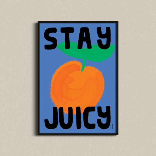 A framed poster featuring a stylized orange on a blue background with the words 'STAY JUICY' in bold black letters, displayed against a light beige textured wall.