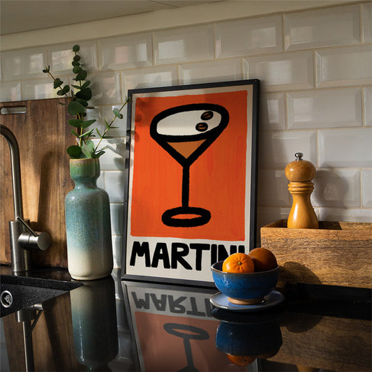 Framed poster with 'MARTINI' design on a kitchen counter with a bowl of oranges and a plant.