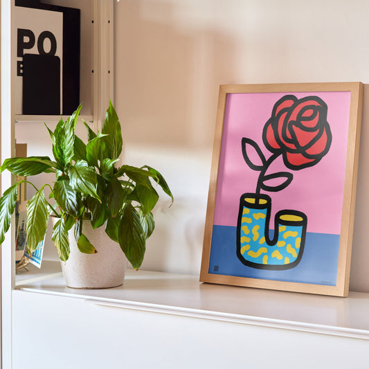 Framed artwork of a rose growing out of boots on a shelf with a plant and lamp.