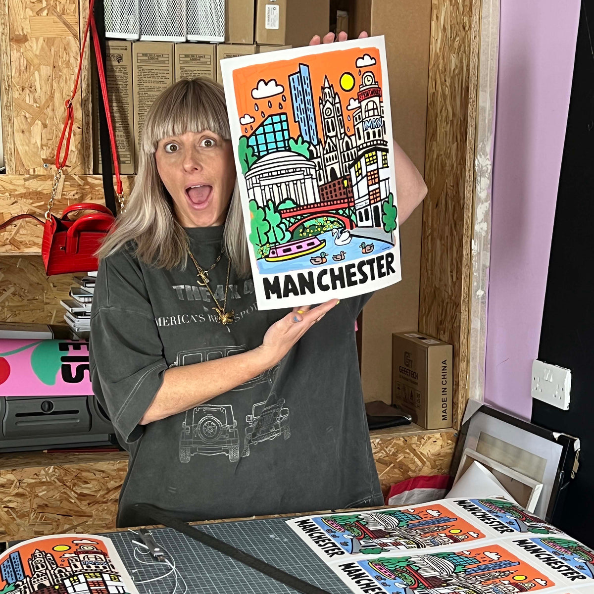 Carla Bentele the artist holds up a colorful Manchester cityscape art print, with more matching prints spread out on a table in front of her. She is wearing a dark grey graphic t-shirt in what appears to be a workshop or studio setting.