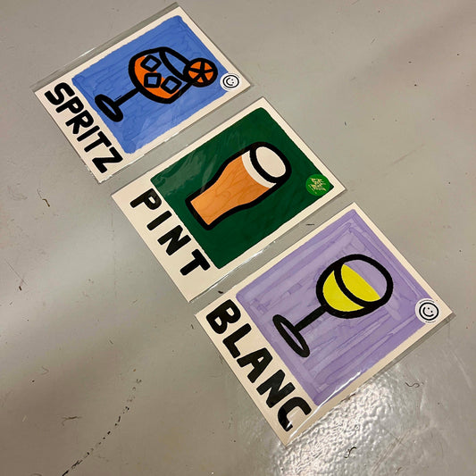 Three small illustrated cards or stickers labeled SPRITZ, PINT, and BLANC, arranged diagonally on a gray surface. Each card shows a simple hand-drawn drink glass icon with a different background color: blue, green, and purple.