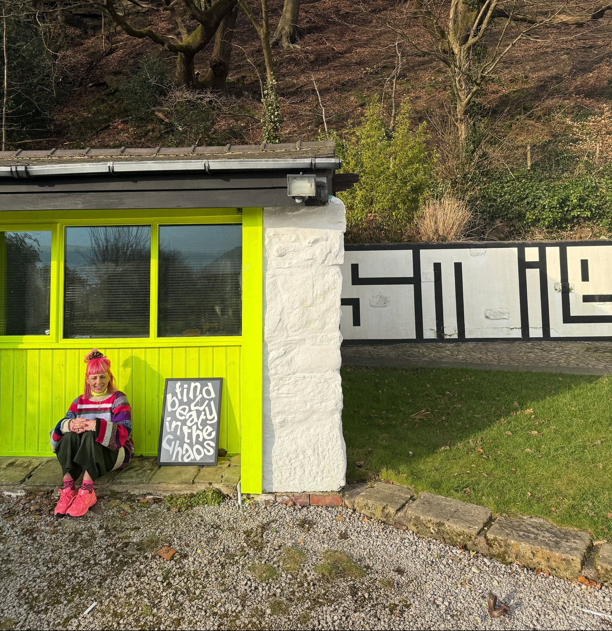 Carla sitting in front of a her bright yellow studio under a canopy of trees  in the background and mural saying smile.