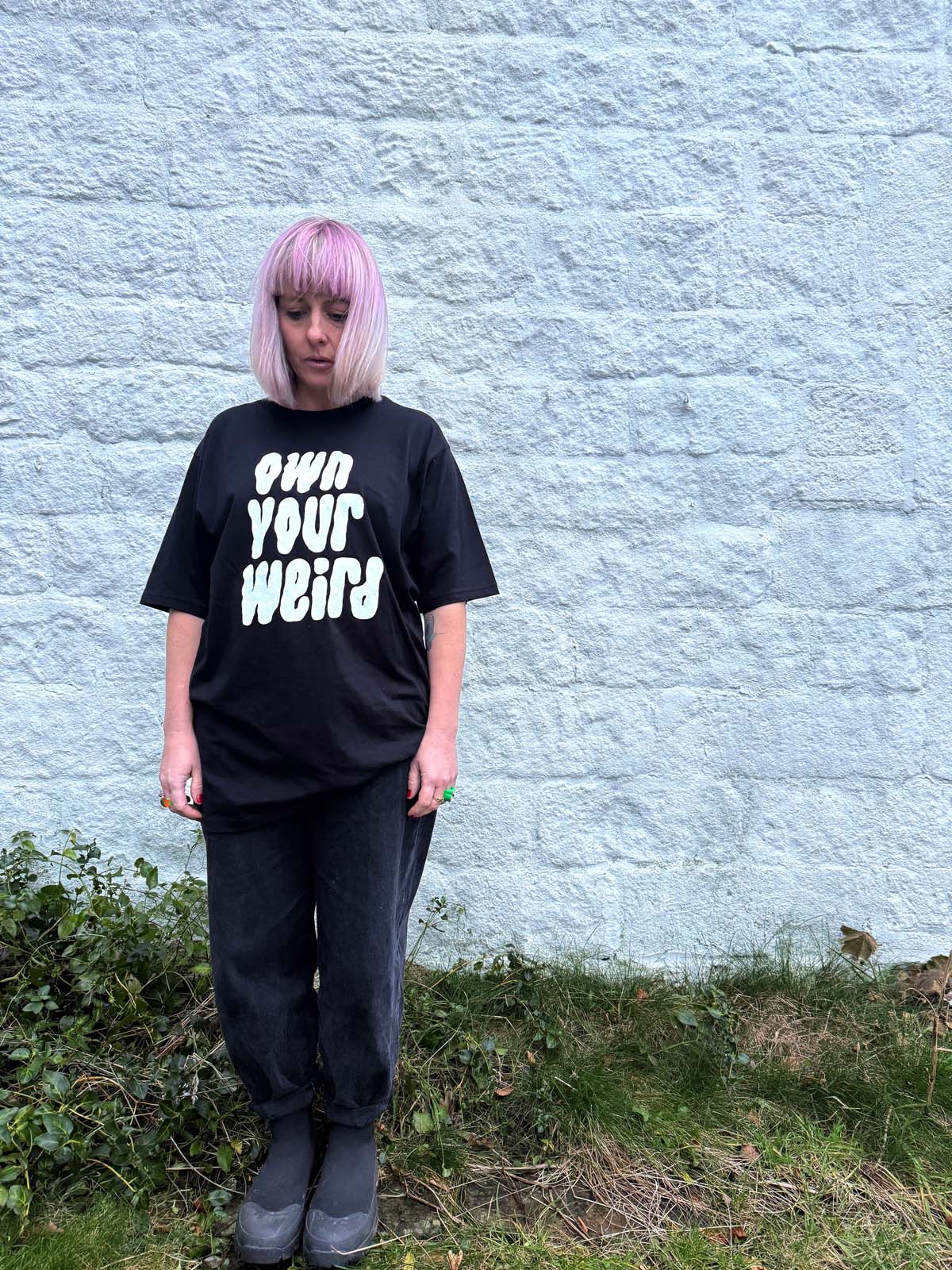 Person wearing a black t-shirt with 'OWN YOUR WEIRD' text against a textured wall.