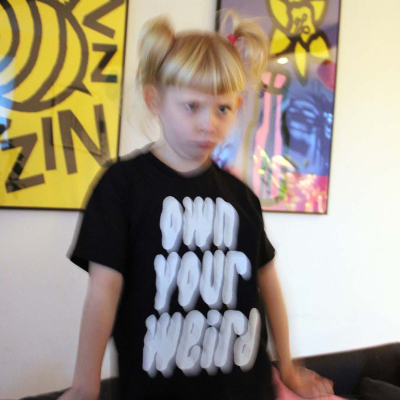 Child wearing a black t-shirt with white text in a room with colorful wall 
art.