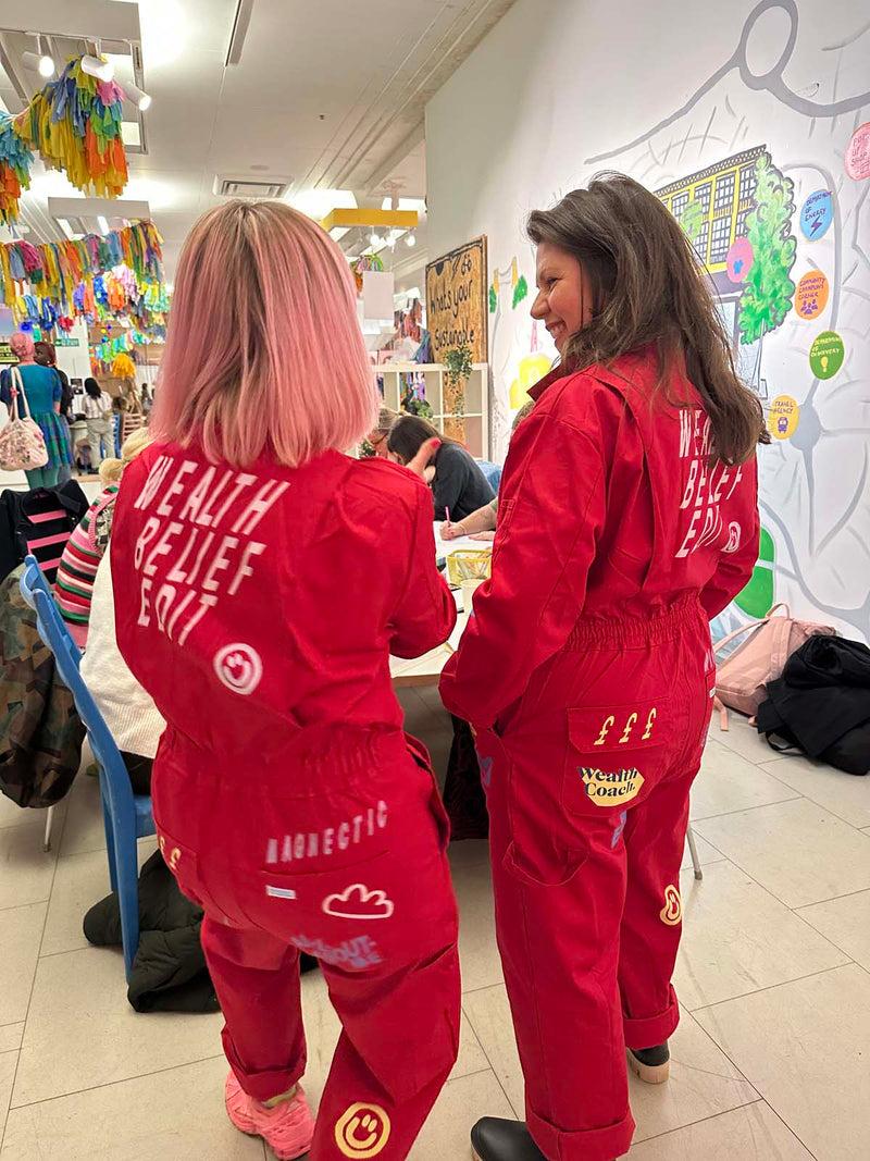 Carla and Sara  wearing red 'wealth belief edit' boilersuits in a workshop.