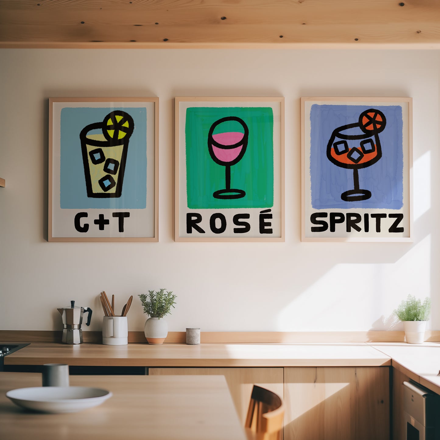 Three framed artworks on a wall above a kitchen counter, each depicting a different type of drink with text underneath.