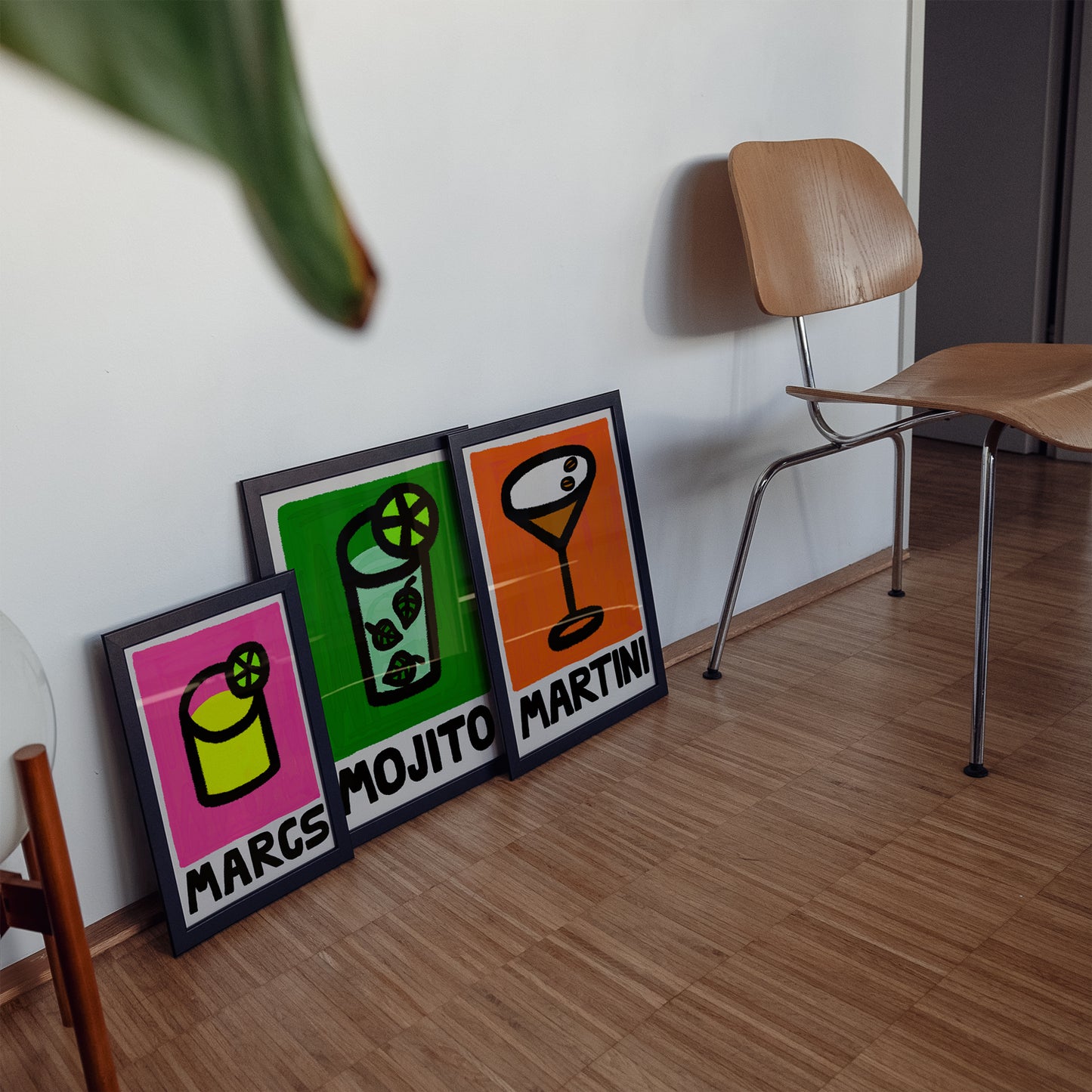 Three colorful framed cocktails posters on a wooden floor with a chair in the background.