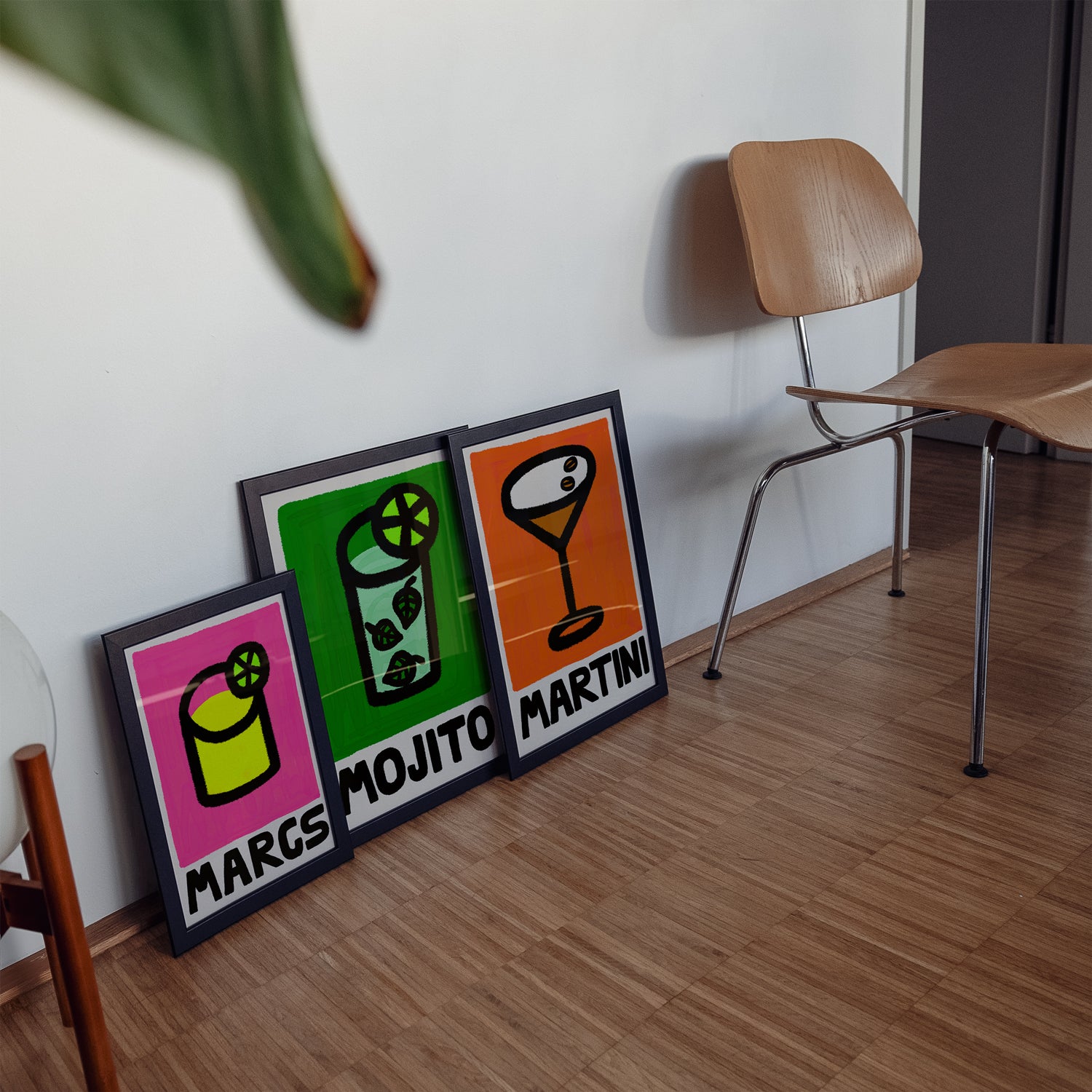 Three colorful framed cocktails posters on a wooden floor with a chair in the background.