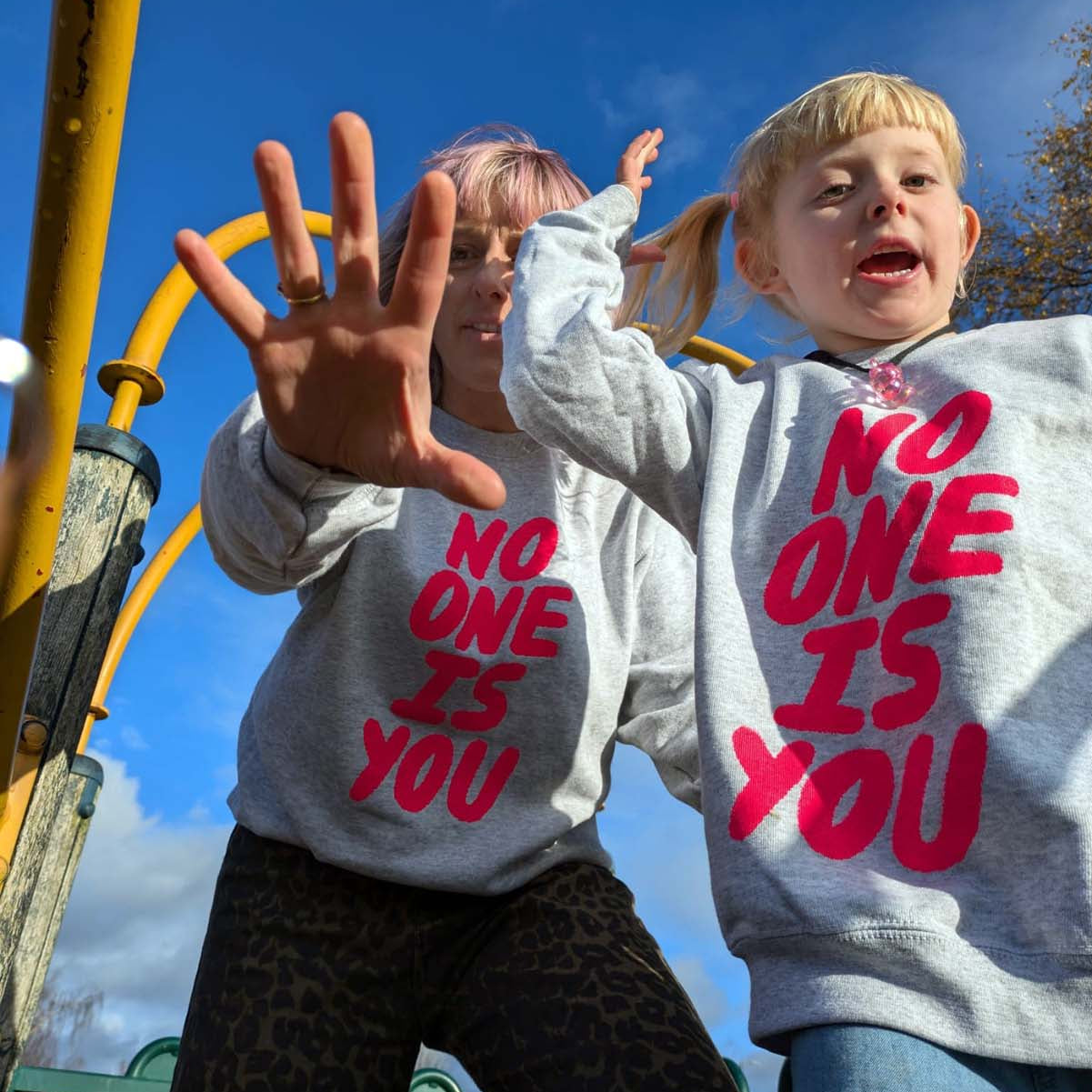 Adult and child wearing sweatshirts with 'NO ONE IS YOU' text on a playground.