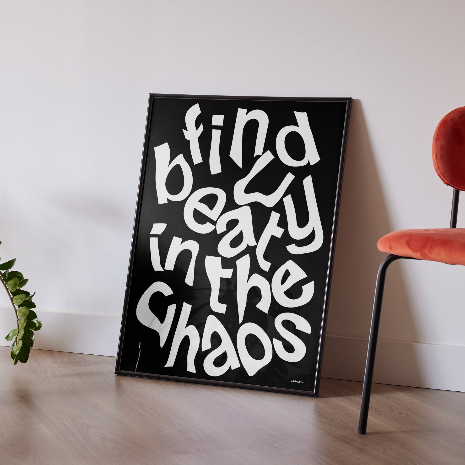 Black framed poster with white text 'Find beauty in the chaos' on a light wooden floor with a red chair and plant.