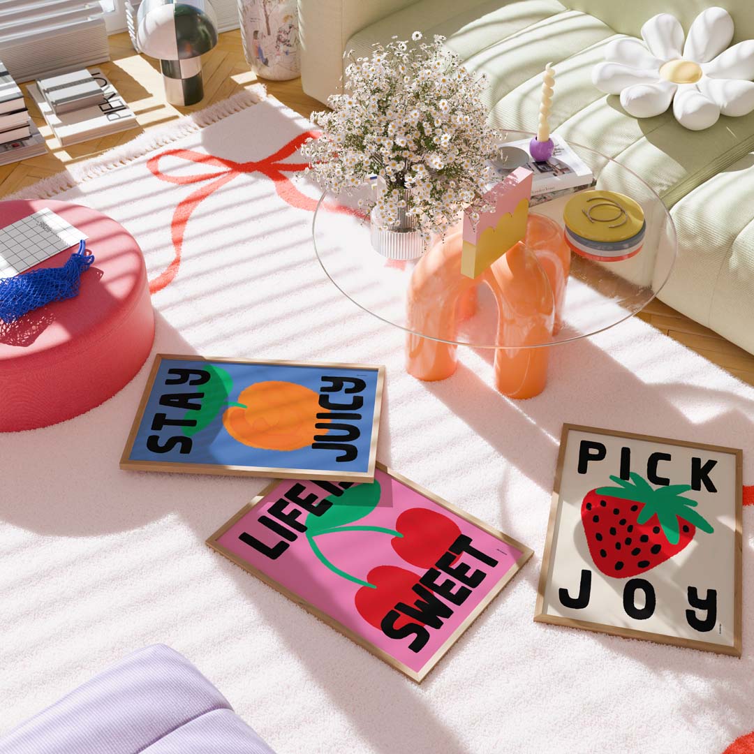 Three decorative rectangular wall art prints with fruit and fruit-themed text are arranged on a white rug in a styled living room. One print says "STAY JUICY" with an orange, one says "LIFE IS SWEET" with cherries, and one says "PICK JOY" with a strawberry.