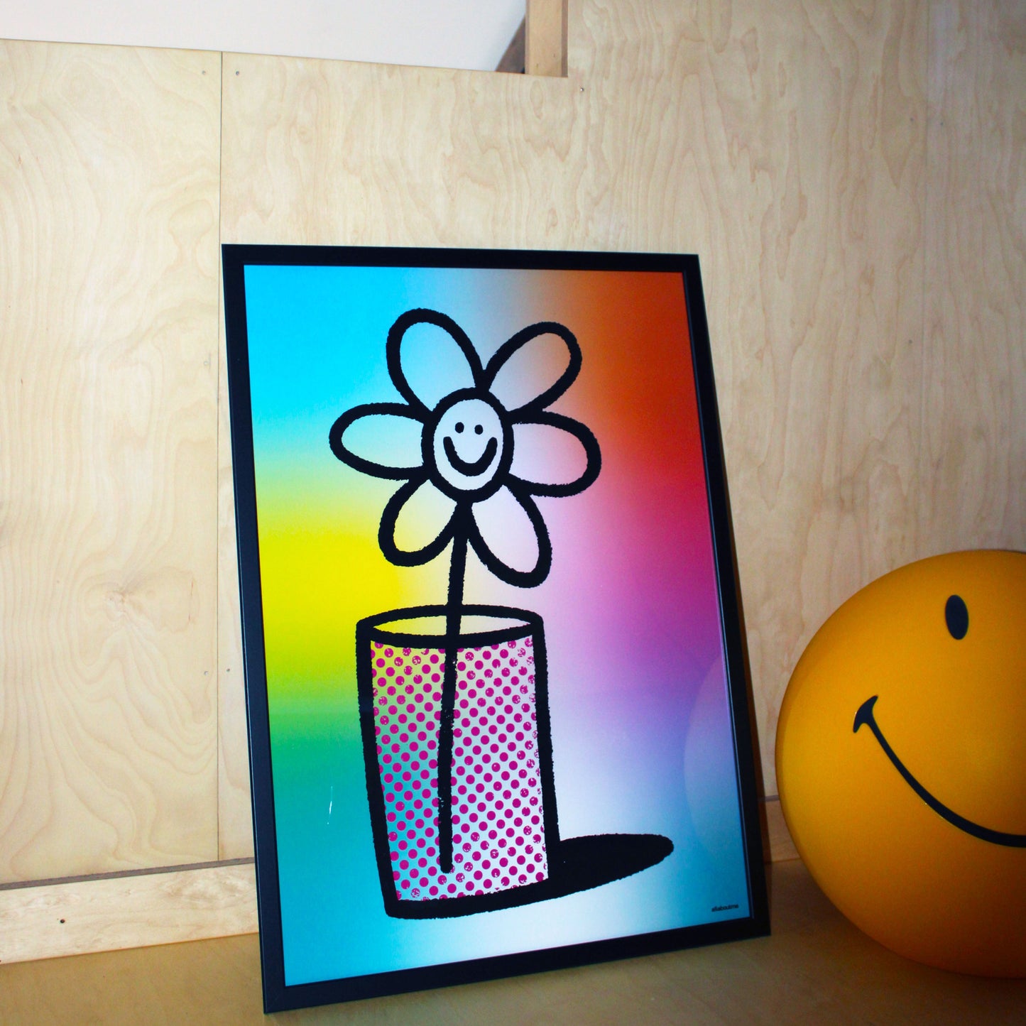 Framed drawing of a flower with a smiley face on a colorful background, leaning against a wooden wall.