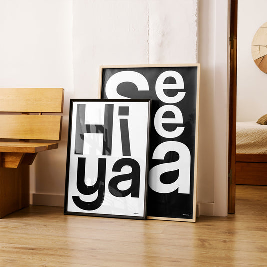 Brighten your space with the Hiya print! Bold, graphic typography art that greets you with positivity and uplifting energy every day.Two framed black and white artworks on a wooden floor with a wooden bench.