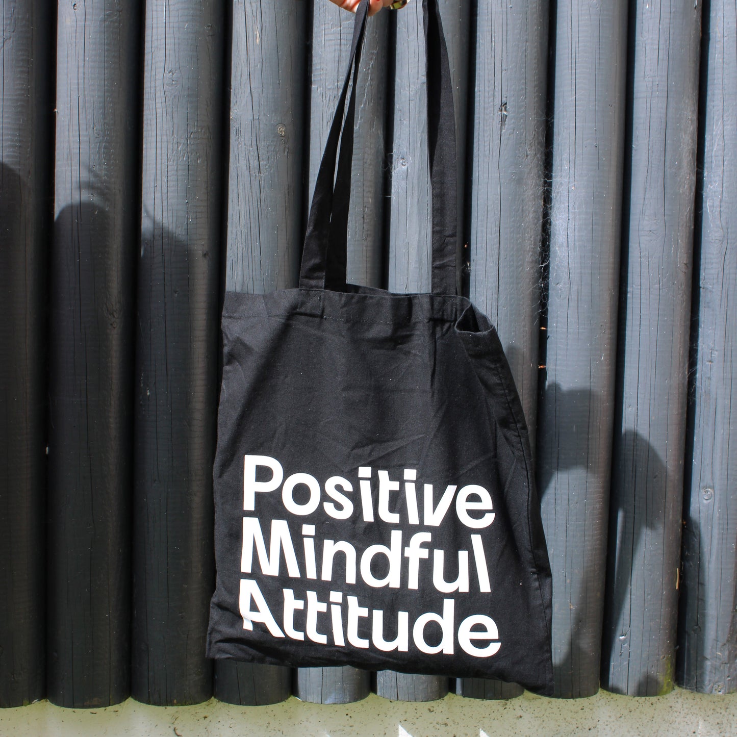 Black tote bag with 'Positive Mindful Attitude' text held by a person against a metallic background