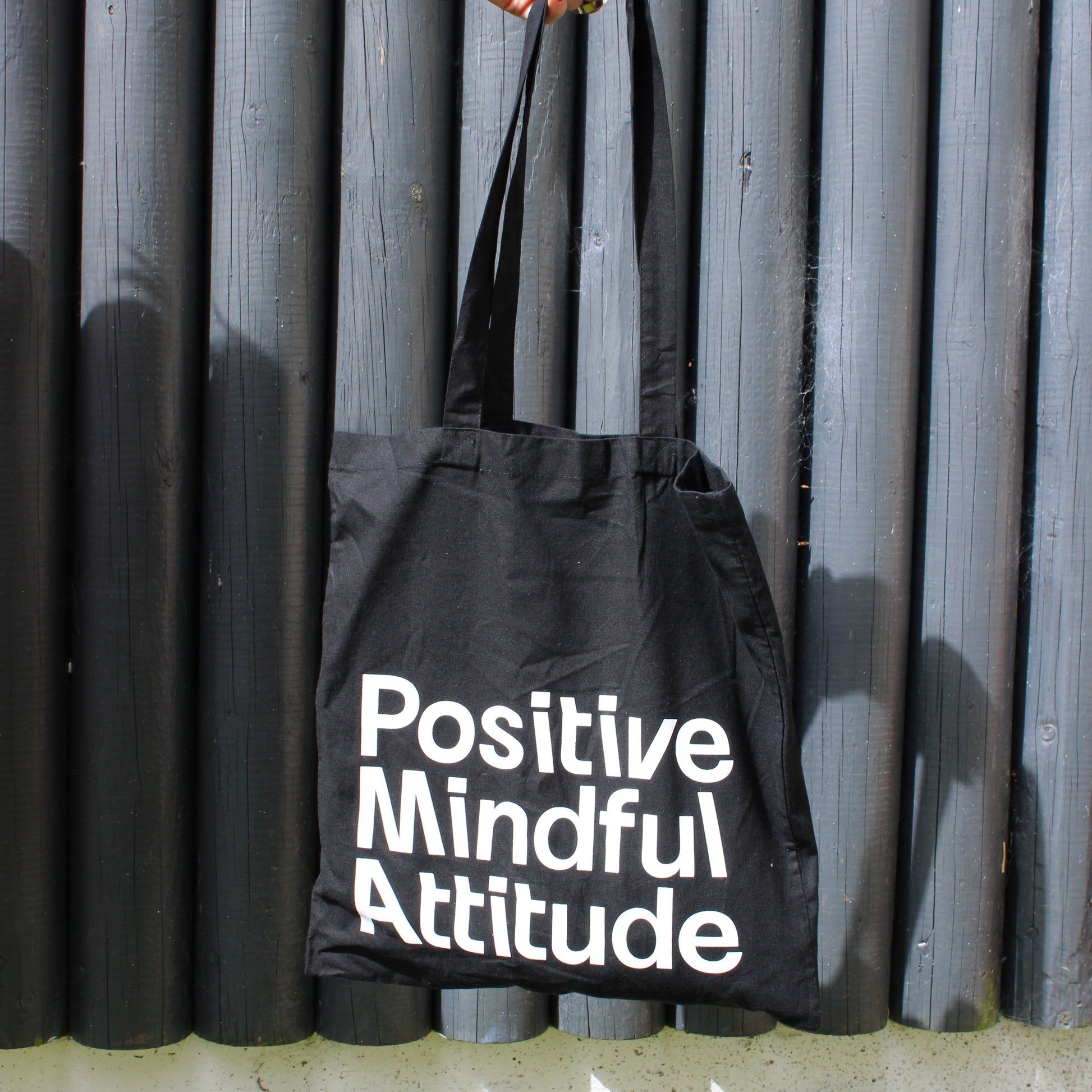 Black tote bag with 'Positive Mindful Attitude' text held by a person against a metallic background