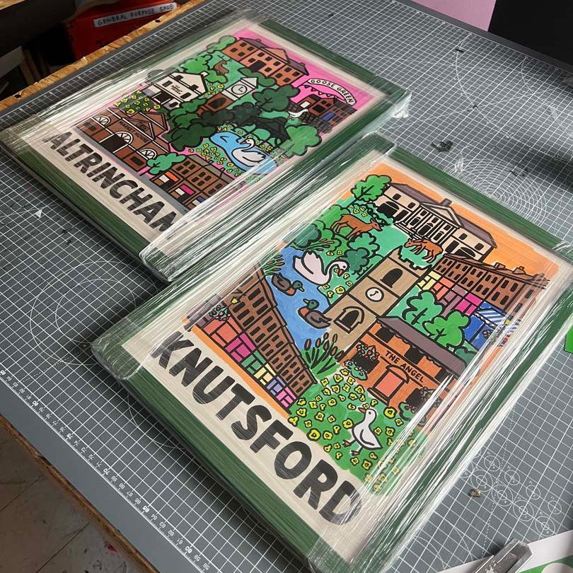 Two framed art prints featuring colorful illustrated town scenes labeled 'ALTRINCHAA' and 'KNUTSFORD' on a gray cutting mat, with a utility knife nearby.