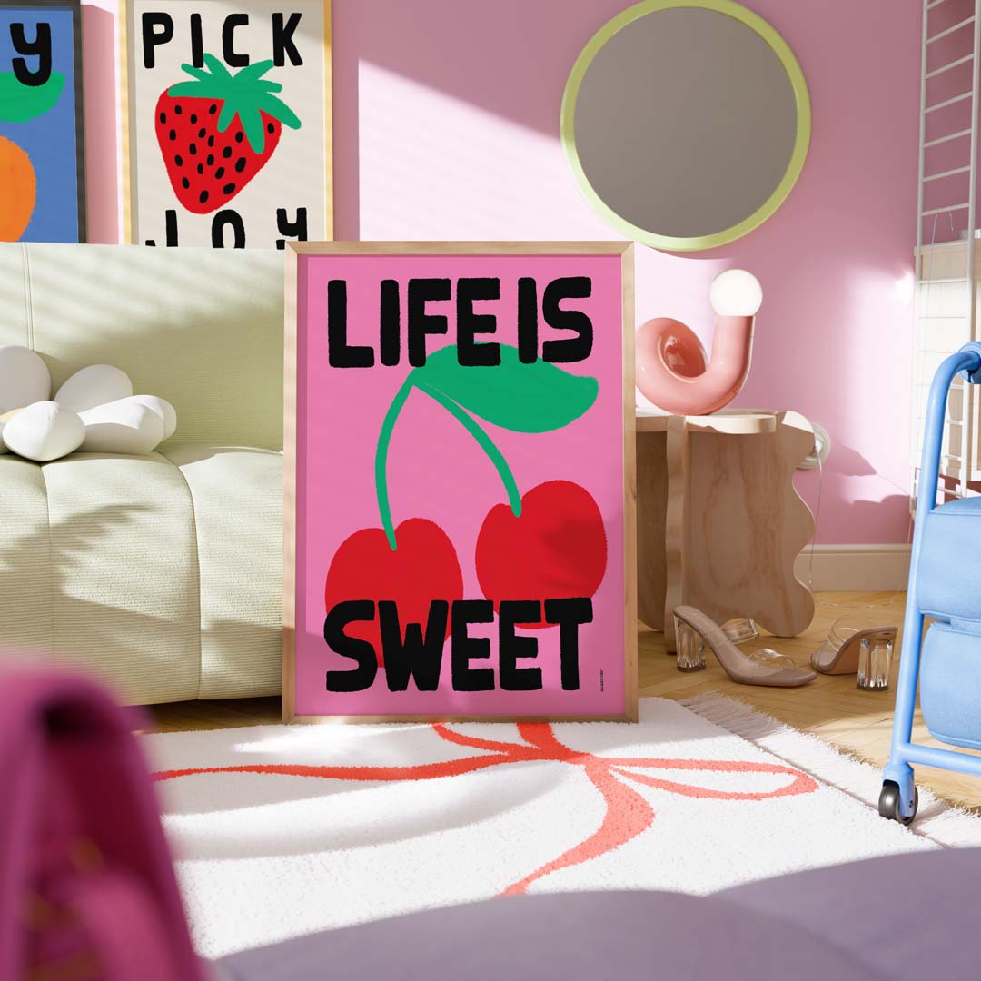 A framed art print with the text 'LIFE IS SWEET' and a pink cherry illustration, displayed in a styled room setting with a pink wall, sofa, and decorative objects.
