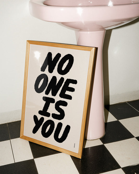 Framed artwork with 'No One Is You' text on a checkered floor with a pink pedestal.