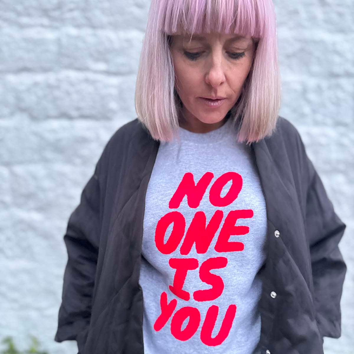 Person wearing a gray sweatshirt with 'NO ONE IS YOU' in neon pink text against a textured wall.