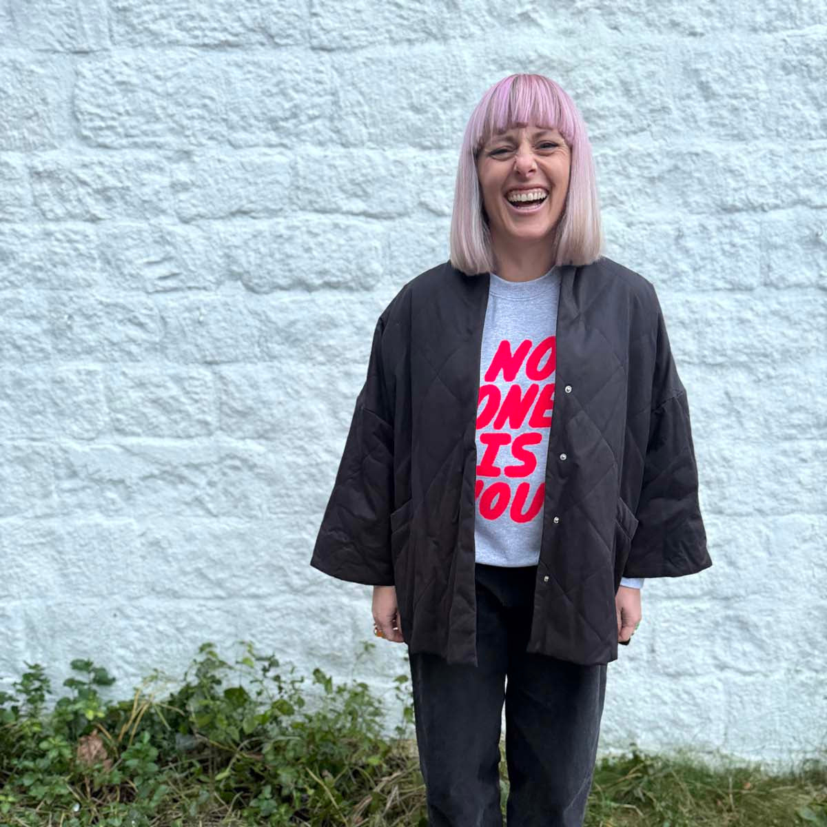 Person wearing a black jacket and neon pink type no one is you empowering sweatshirt standing against a light gray wall.
