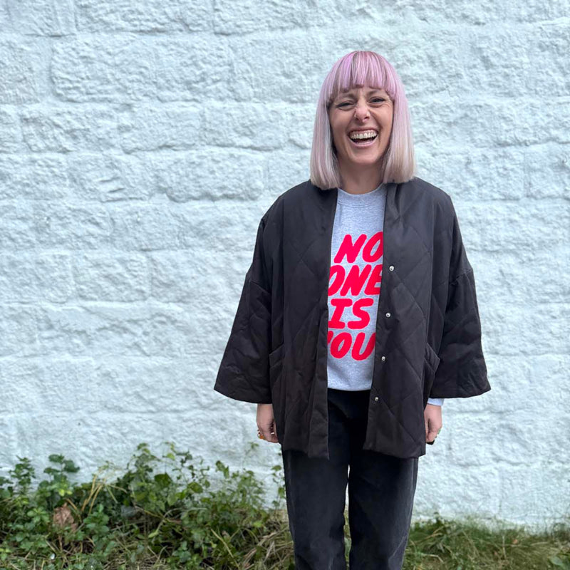 Person wearing a black jacket and neon pink type no one is you empowering sweatshirt standing against a light gray wall.