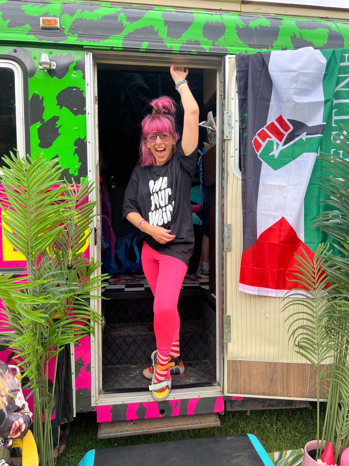 Person with pink hair and colorful outfit standing in the doorway of a vibrant, decorated vehicle.
