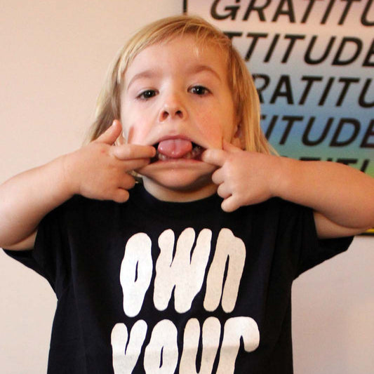 Child wearing a black t-shirt with 'OWN YOUR WEIRD' text, making a playful gesture.