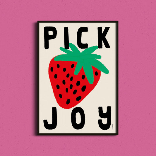 A framed art print featuring a bold graphic design with the text 'PICK JOY' and a large red strawberry illustration, displayed against a solid pink background.