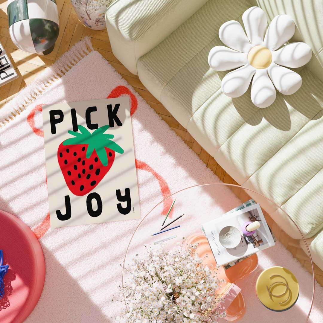 A styled flat-lay featuring a rectangular cream card with a large red strawberry graphic and the words 'PICK JOY' in black, placed on a pink rug beside a pale green sofa, a white flower-shaped pillow, and a glass table with small accessories.