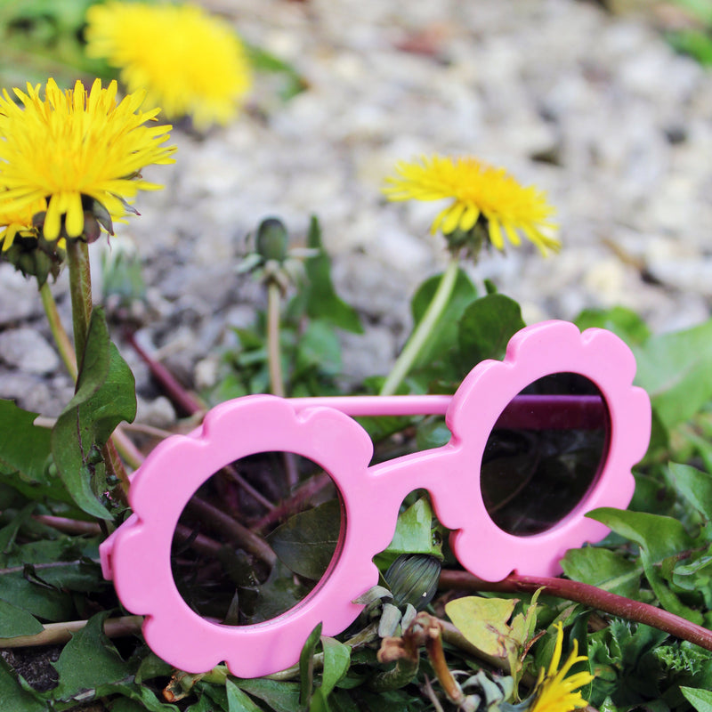 Pink sunglasses with floral lenses on a background of green leaves and yellow flowers.