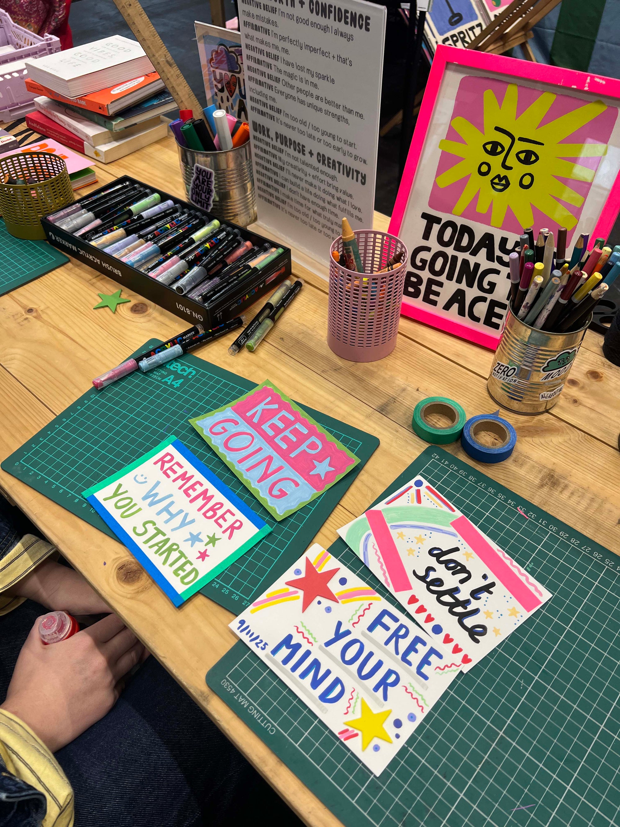 Table with art supplies, motivational posters, and a person's hand on a green cutting mat.