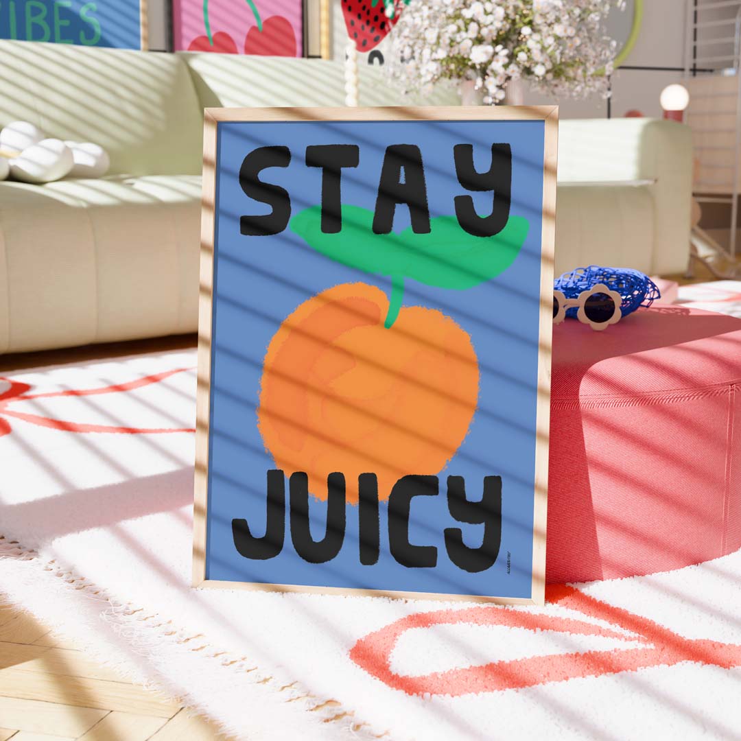 A framed art print with the text 'STAY JUICY' and a large orange illustration on a blue background, styled in a brightly lit living room setting.