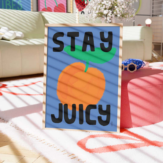 A framed art print with the text 'STAY JUICY' and a large orange illustration on a blue background, styled in a brightly lit living room setting.