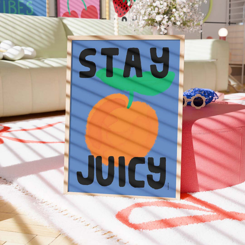 A framed art print with the text 'STAY JUICY' and a large orange illustration on a blue background, styled in a brightly lit living room setting.