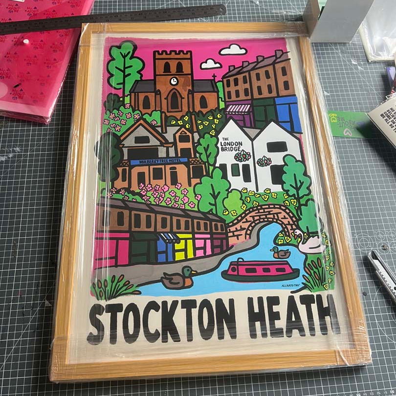 A framed hand-drawn illustration of Stockton Heath featuring colorful houses, trees, a canal, ducks, and the text ' STOCKTON HEATH' at the bottom, photographed on a cutting mat with art supplies around it.