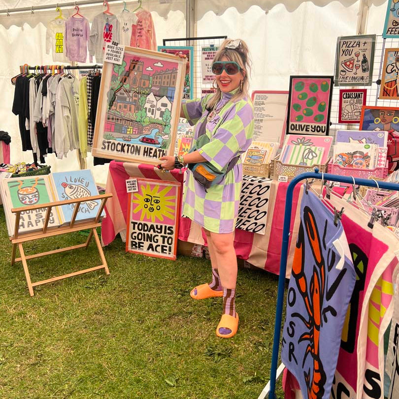 Carla wearing a pastel checkered shirt, shorts, and sunglasses stands under a white market tent holding a colorful art print. The scene shows a stokton heah festival with clothing racks, stickers, and other art pieces on a grassy floor.
