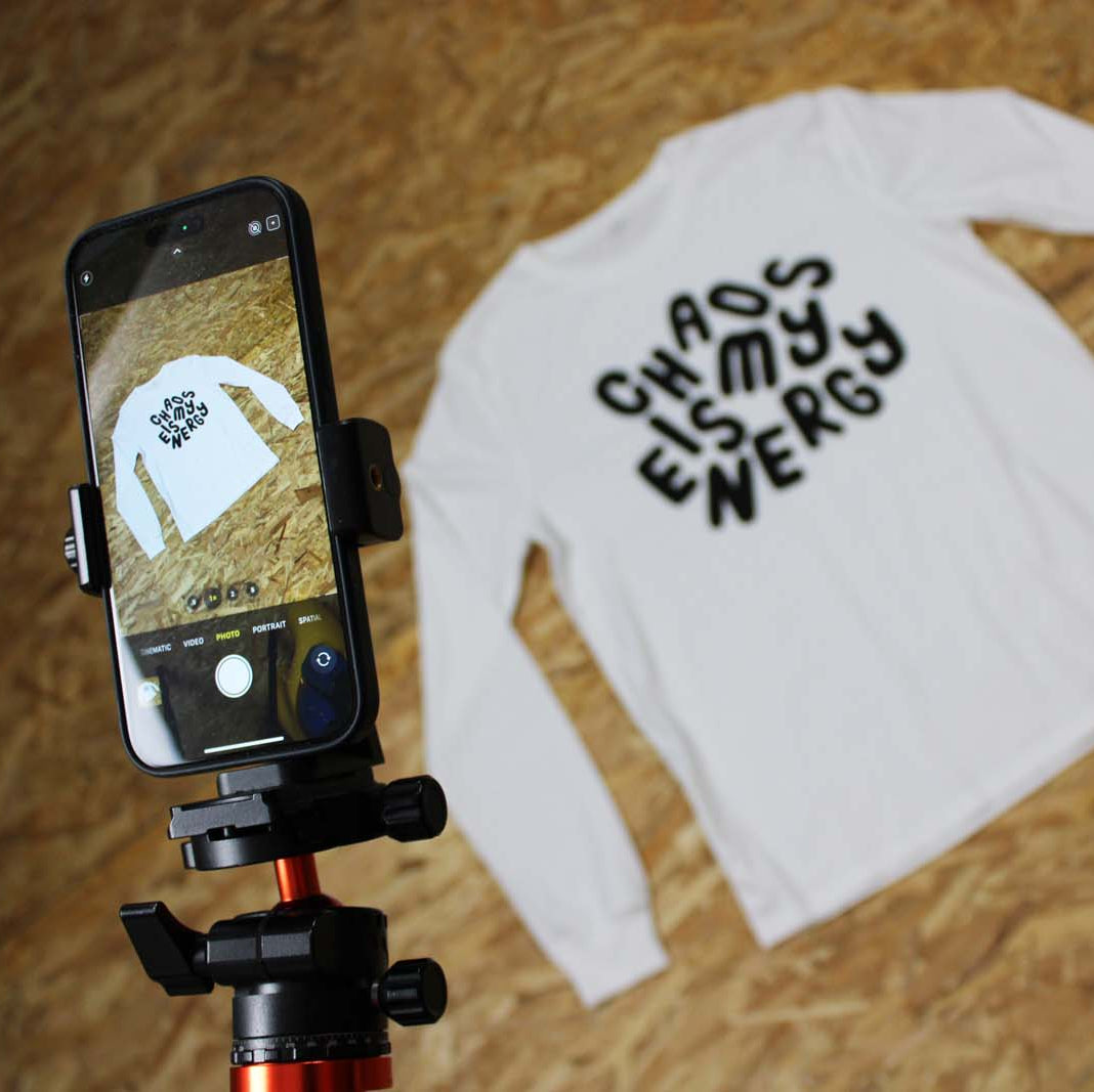 White long-sleeve shirt with 'CHAOS IS MY ENERGY' text on a wooden floor, with a phone.