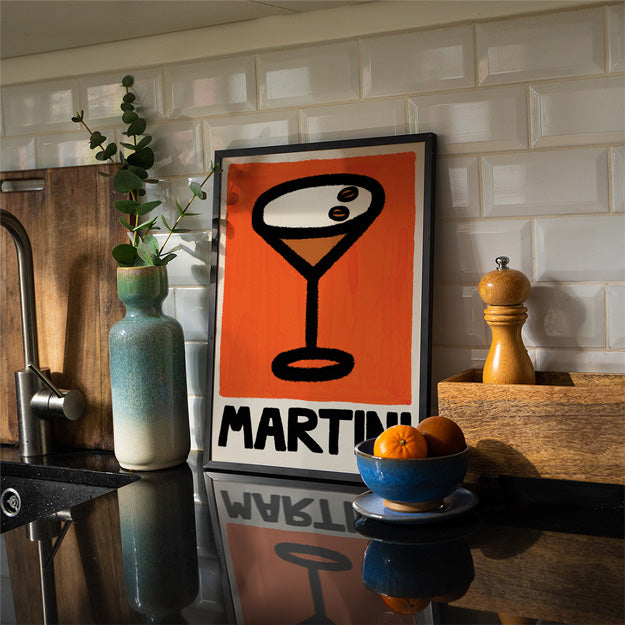 Framed poster with 'MARTINI' design on a kitchen counter with a bowl of oranges and a plant.