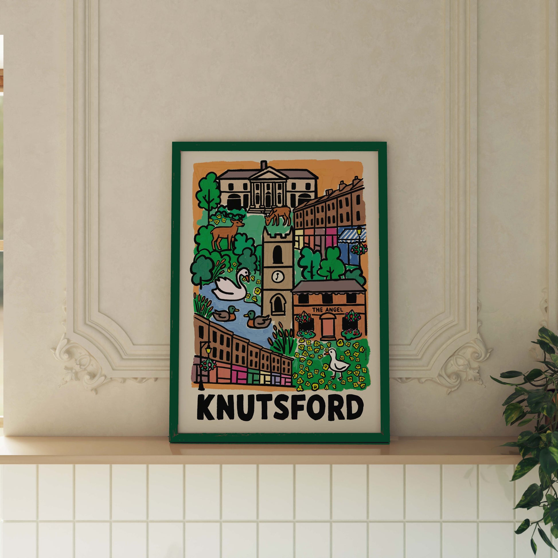 A framed art print featuring a colorful illustrated town scene labeled 'KNUTSFORD' at the bottom, displayed on a light wood mantel against a cream wall with decorative molding and a potted plant at the side.