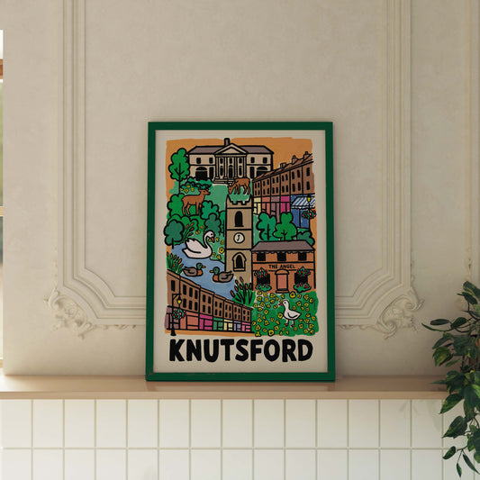 A framed art print featuring a colorful illustrated town scene labeled 'KNUTSFORD' at the bottom, displayed on a light wood mantel against a cream wall with decorative molding and a potted plant at the side.