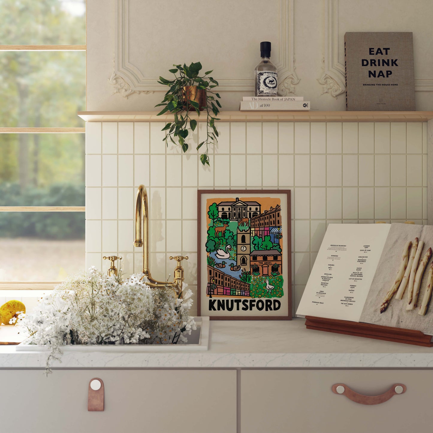 A framed travel-style poster reading 'KNUTSFORD' hangs on a kitchen wall above a sink. The scene includes a gold faucet, white flowers in the sink, an open book, a small plant, and a book titled 'EAT DRINK NAP'.