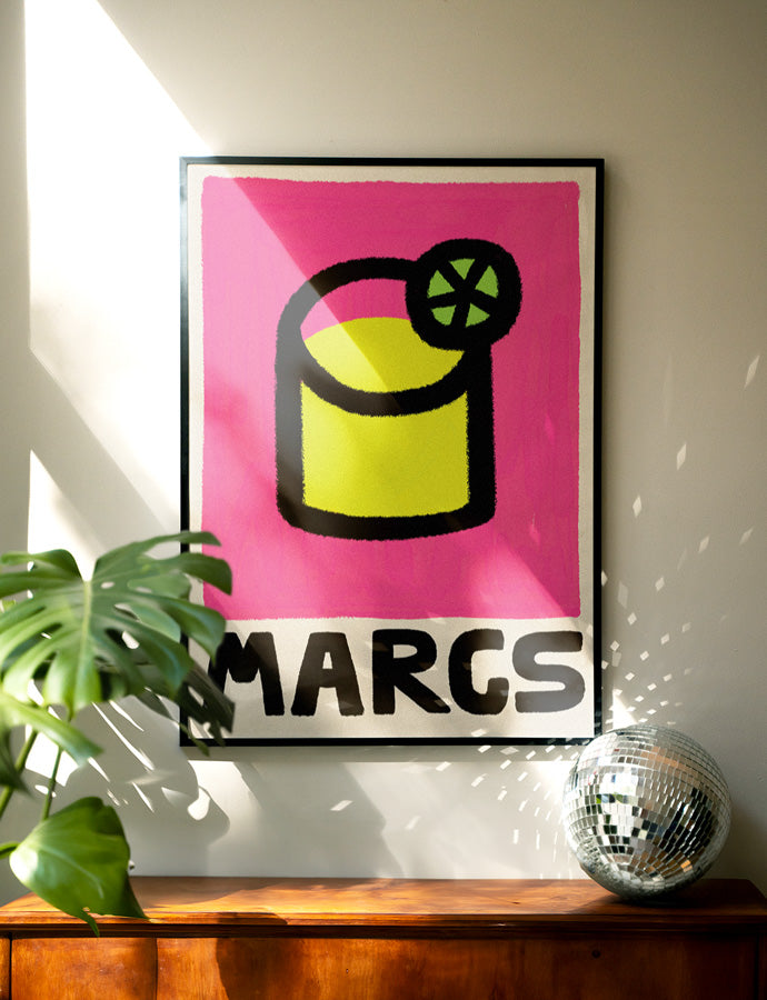Framed poster with a colorful design of margarita cocktail and 'MARGS' text on a wall with a plant and disco ball.