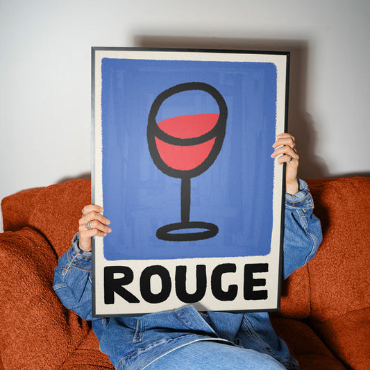 Person holding a framed poster with a wine glass and 'ROUGE' text against a white wall.