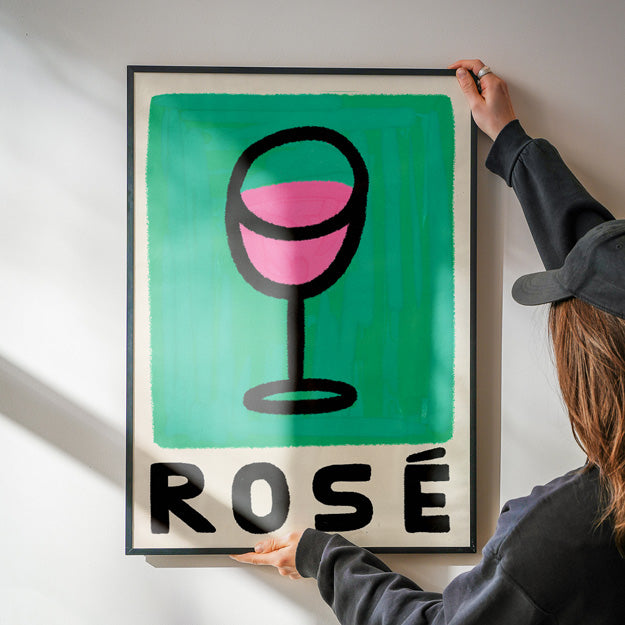 Person holding a framed poster with a wine glass and 'Rosé' text against a white wall.