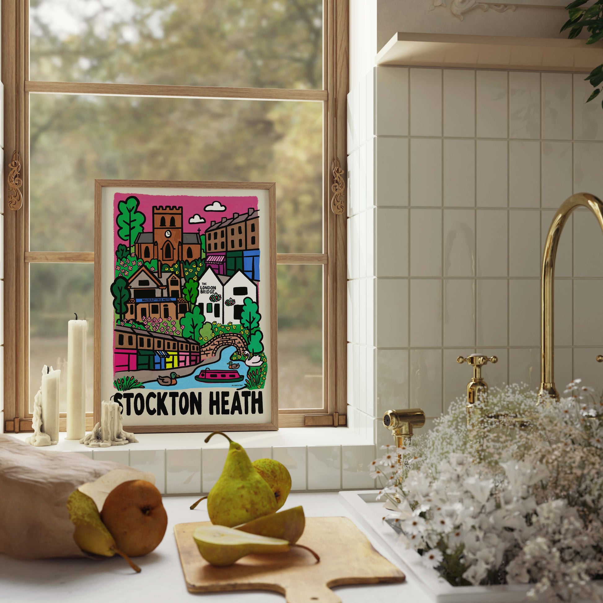 A colorful framed art print featuring an illustration of Stockton Heath with the text 'STOCKTON HEATH' at the bottom, displayed on a kitchen windowsill next to candles, pears, and a sink with white flowers.